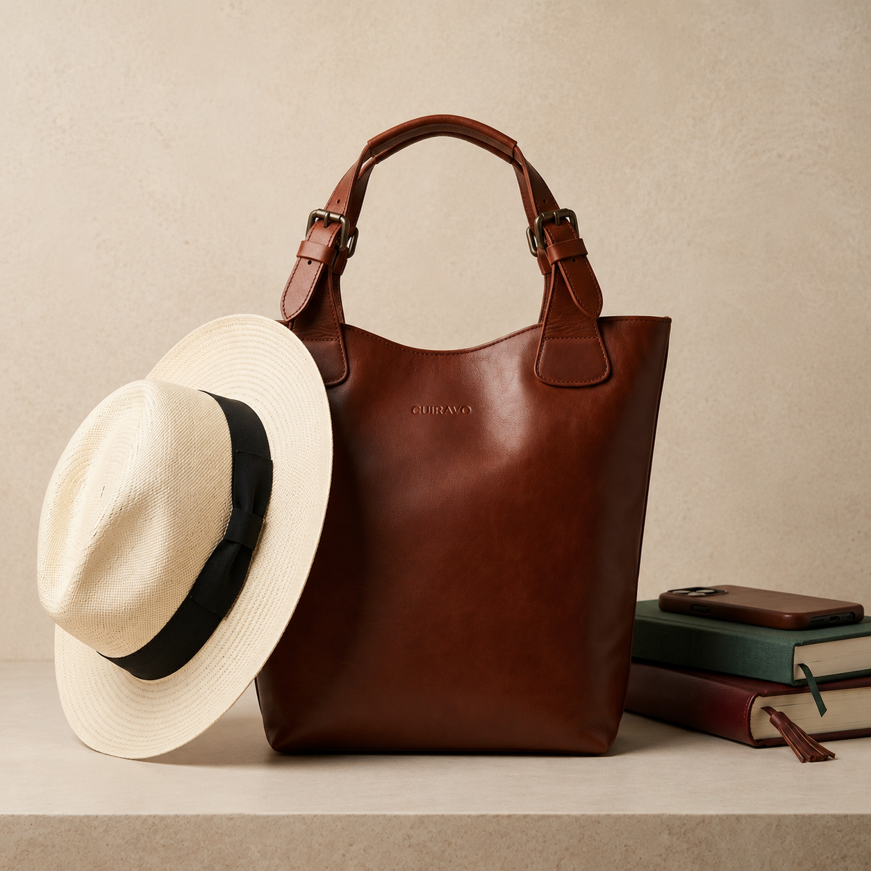 A brown leather Cuiravo Amara tote bag stands on a light stone surface with a cream straw hat and stacked books beside it. The bag has top handles, buckle details, and a small embossed brand mark that appears to read Cuiravo.