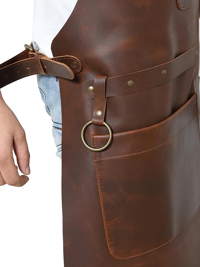 Close-up of brown leather apron side pocket, brass tool ring, rivets, and adjustable waist strap