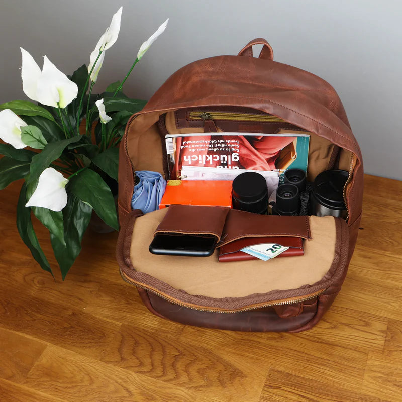 Open brown Atlas leather backpack packed with reading materials, phone, wallet, and small travel essentials