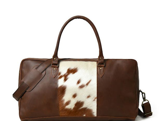 A brown leather duffel bag with a brown and white cowhide panel on the front, featuring two top handles and a detachable shoulder strap, set against a plain white background.