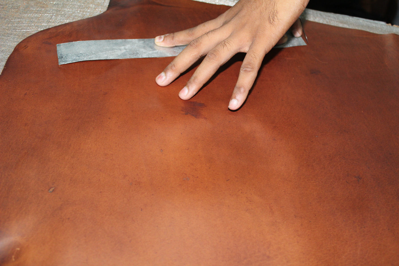 A close-up of a person's hand holding a metal ruler against a large, smooth, reddish-brown leather surface, likely showing the grain or thickness of the material.