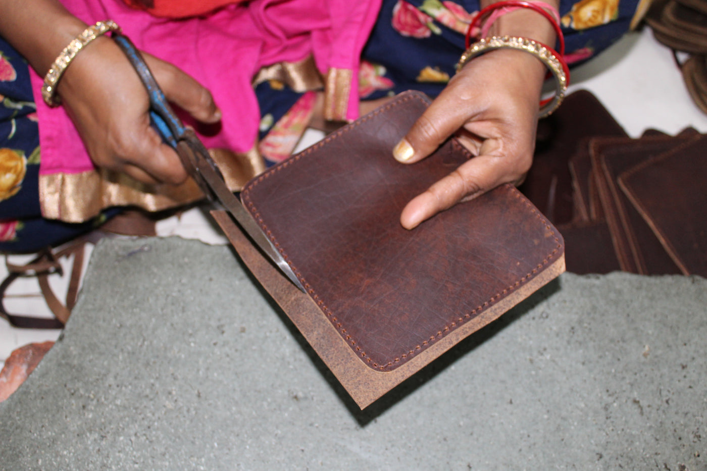 A close-up of a person's hands cutting a brown leather patch cover with blue-handled scissors, wearing a bright pink and floral traditional outfit.