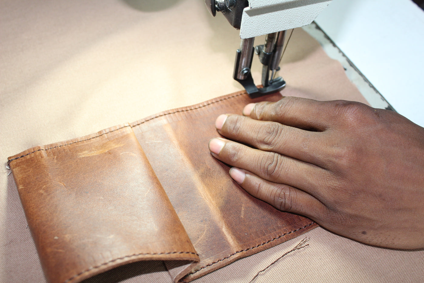 A close-up of a brown leather patch being sewn by a hand using a sewing machine, with a beige fabric background.