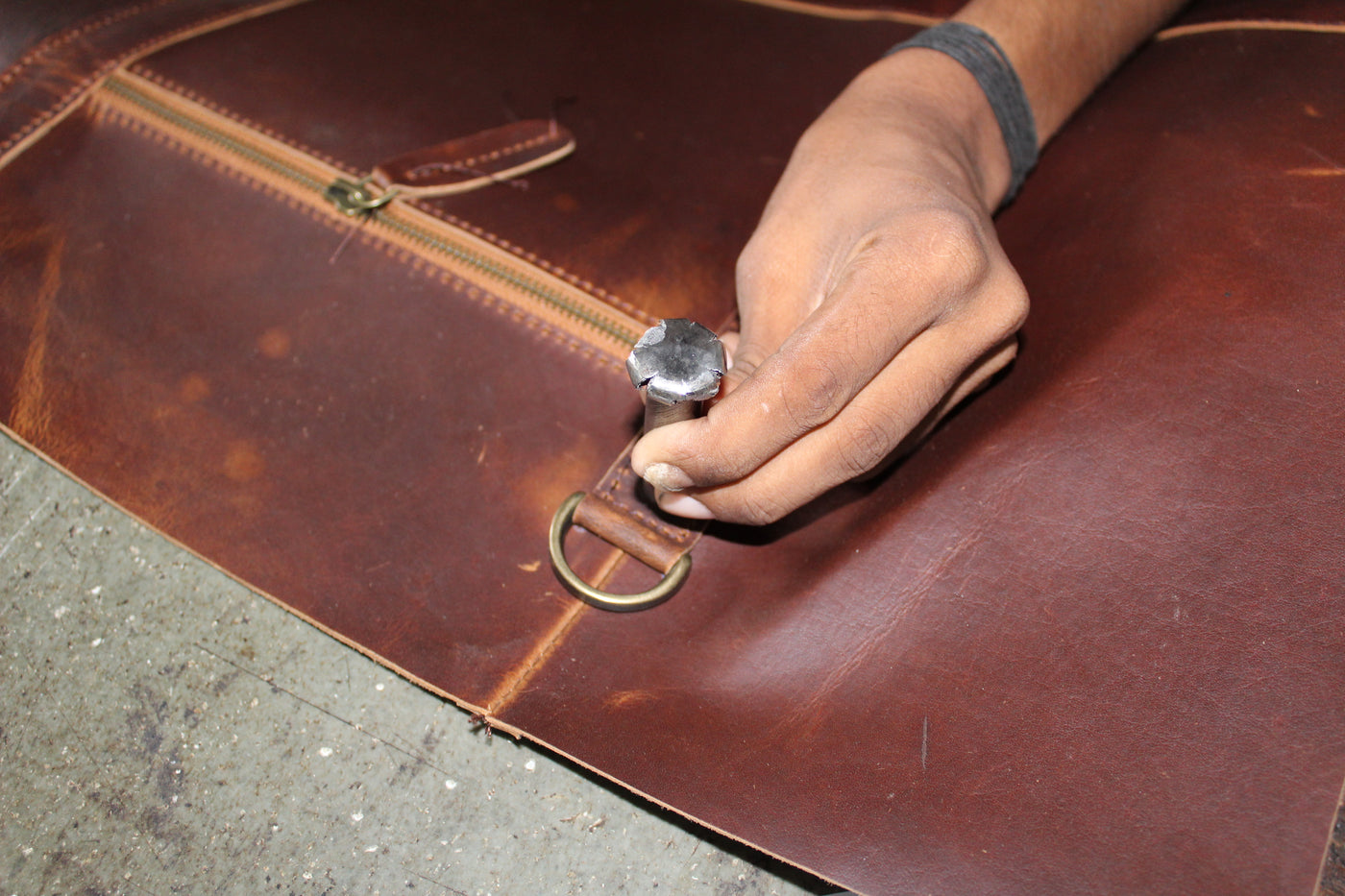 A close-up of a person's hand using a leather punch tool to create a leather stitch on a brown leather bag.