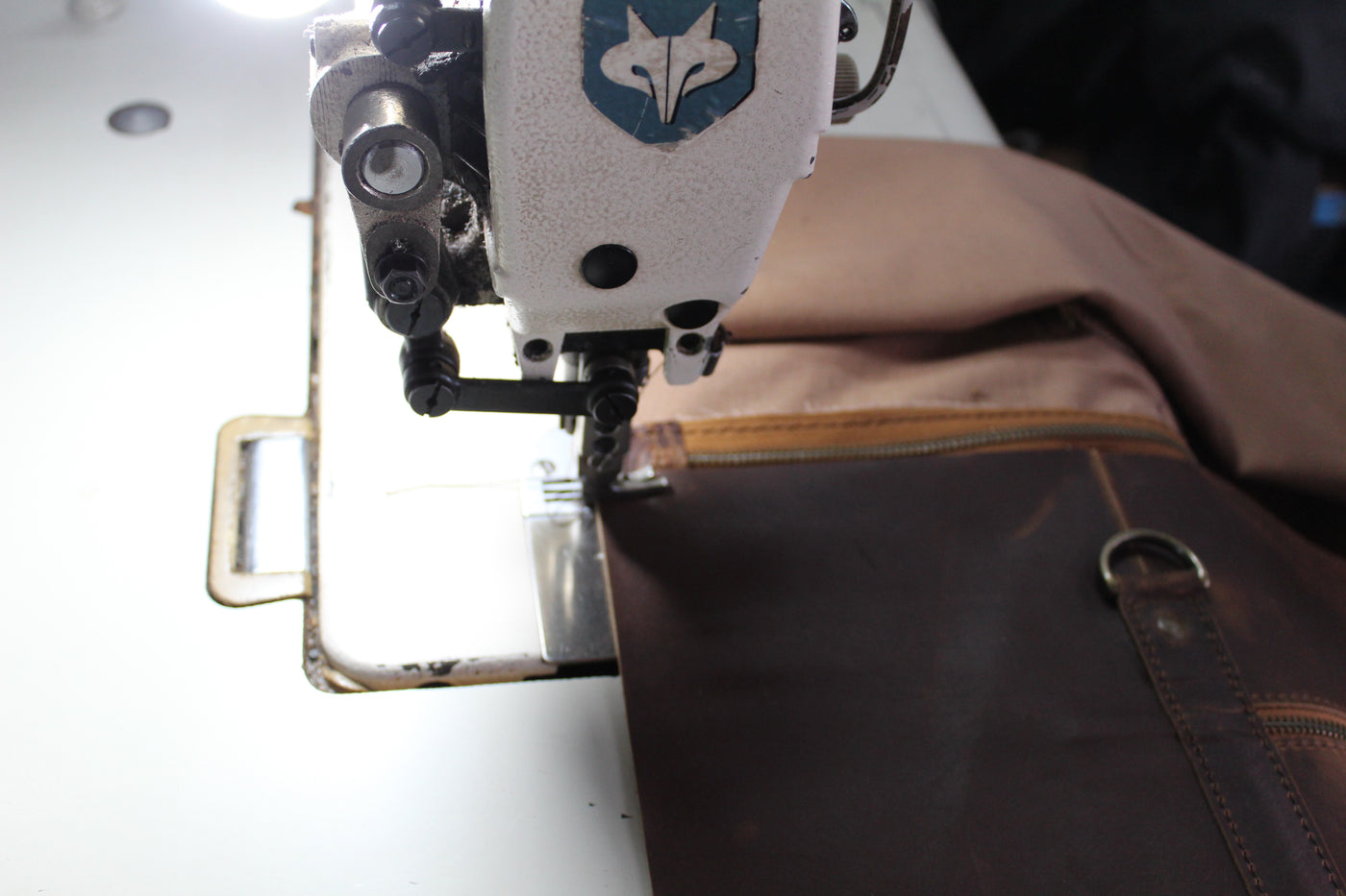A close-up of a sewing machine stitching a dark brown leather apron or pouch next to a tan fabric piece on a white sewing table.