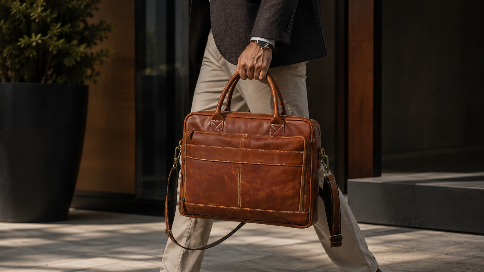 A man in a dark blazer and beige trousers carries a brown Cuiravo Rivelle leather briefcase with a top handle and shoulder strap in an outdoor urban setting.